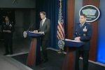 A man in a suit and a man in uniform stand at podiums in front of a Pentagon sign.