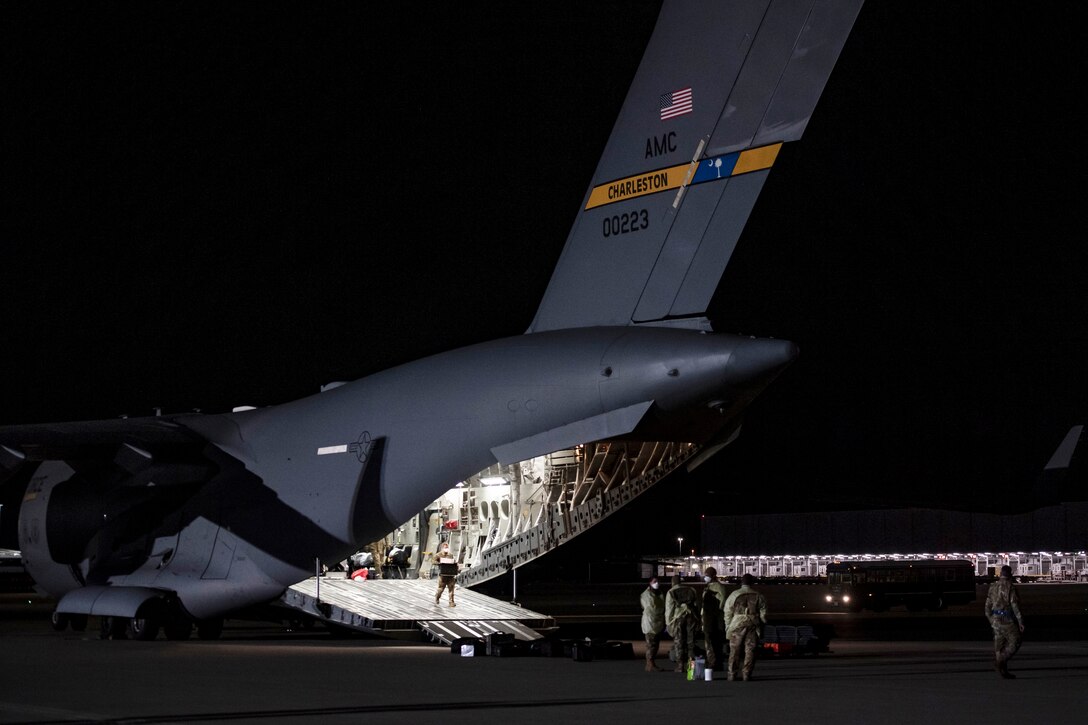 Airmen remove equipment from the brightly lit cargo bay of a large transport jet on a dark flight line.