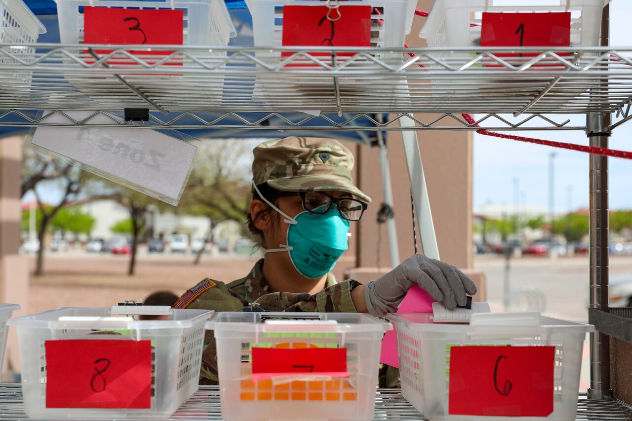 A soldier wearing a face mask works at a shelving system outside containing numbered bins.