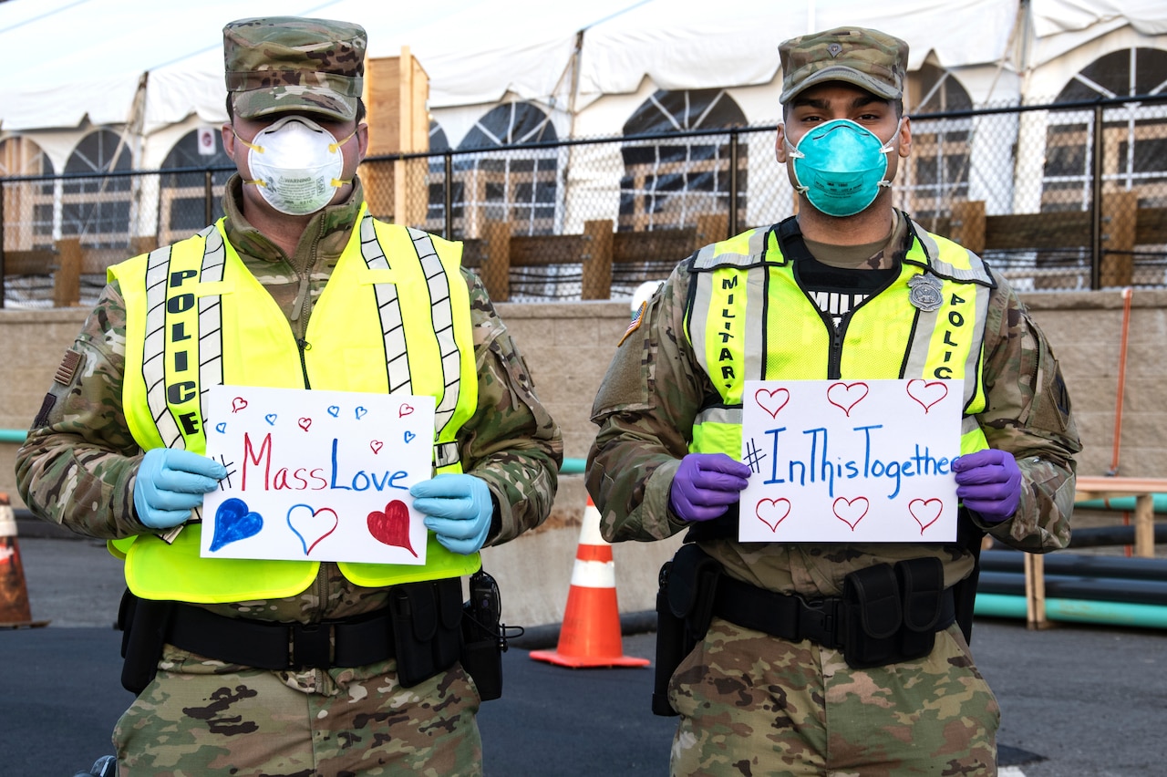 Service members wearing face masks hold signs that read “#MassLOve” and “#In This Together.”