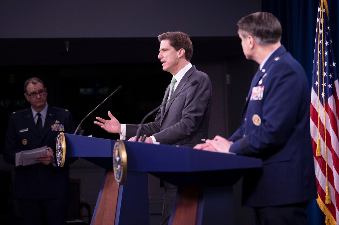 A civilian speaks at a lectern as an Air Force general stands beside him at an identical lectern.