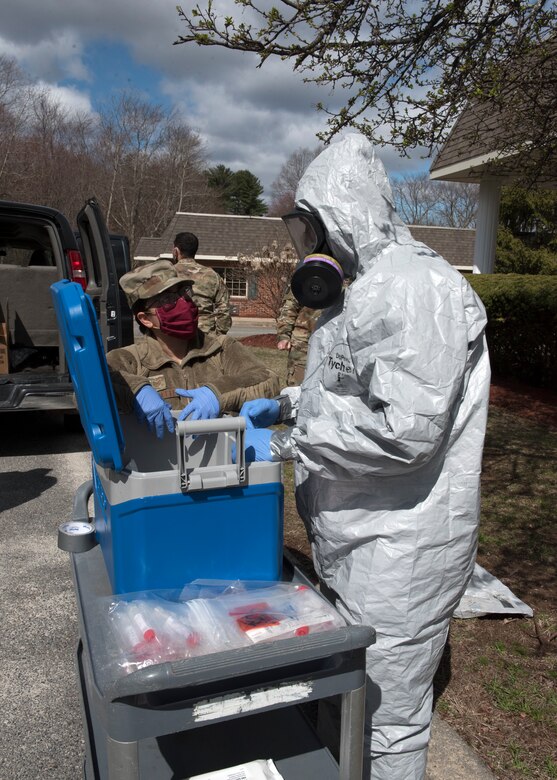 Two service members wearing personal protective equipment talk as they stand by a cart bearing a blue cooler and medical equipment.