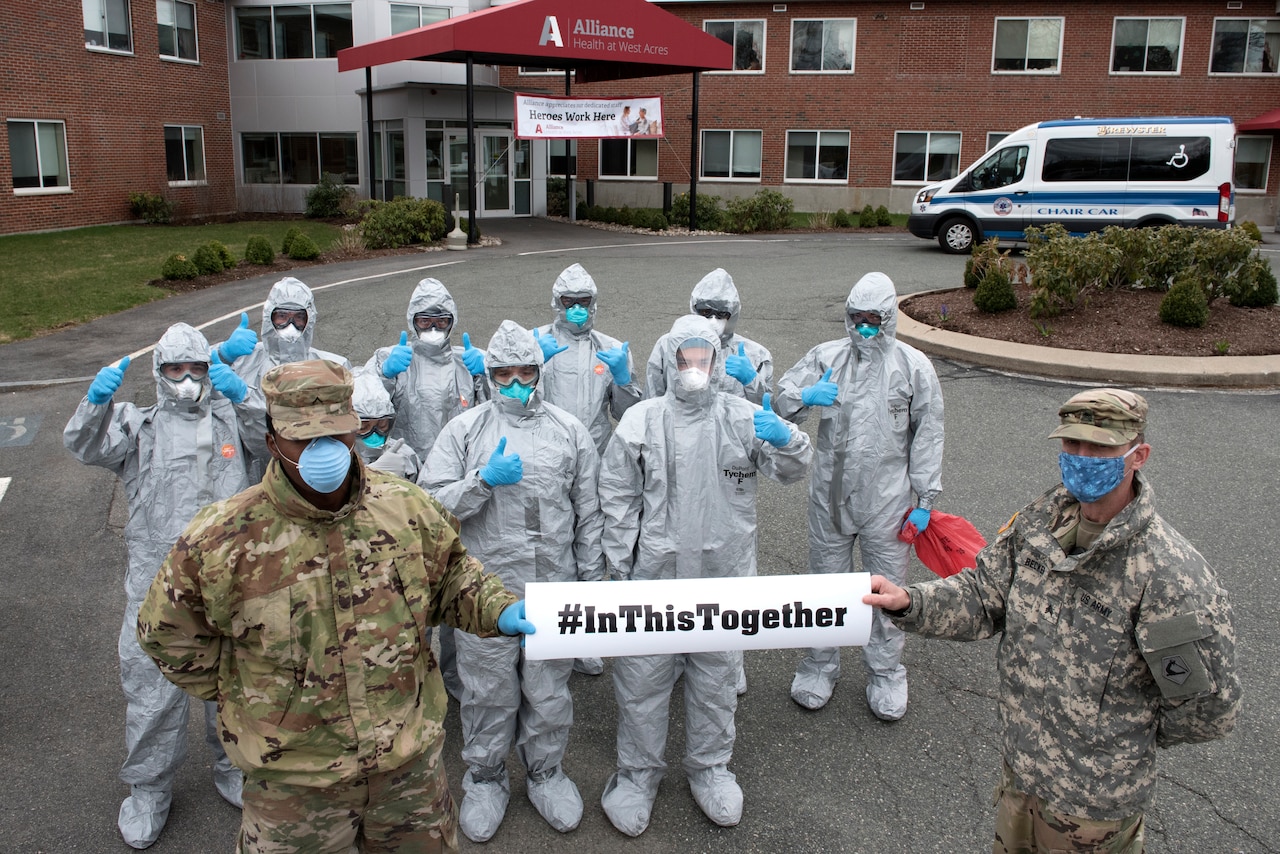 Service members wearing personal protective equipment pose in front of a nursing home holding a sign that reads, “#In This Together.”