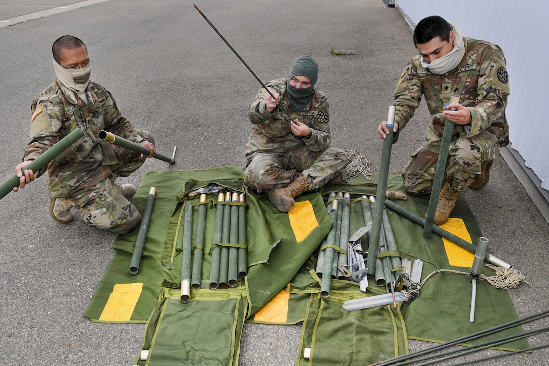 Three soldiers in facemasks assemble an antenna outdoors.