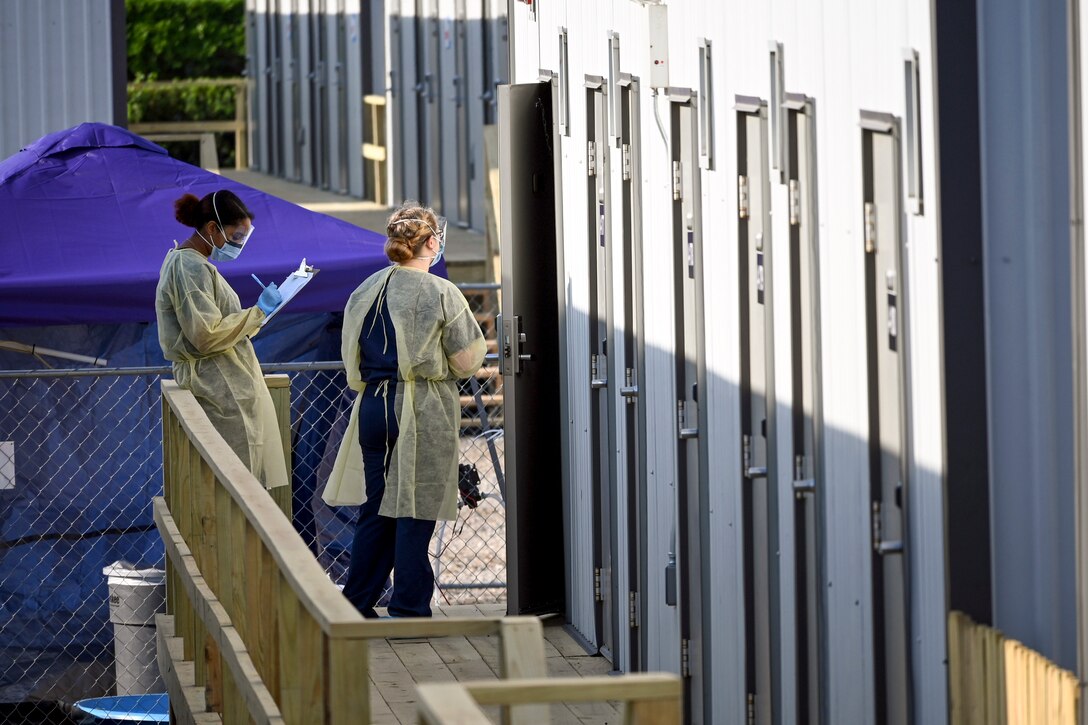 Two sailors in protective medical gear stand outside an ajar front door at a housing facility.