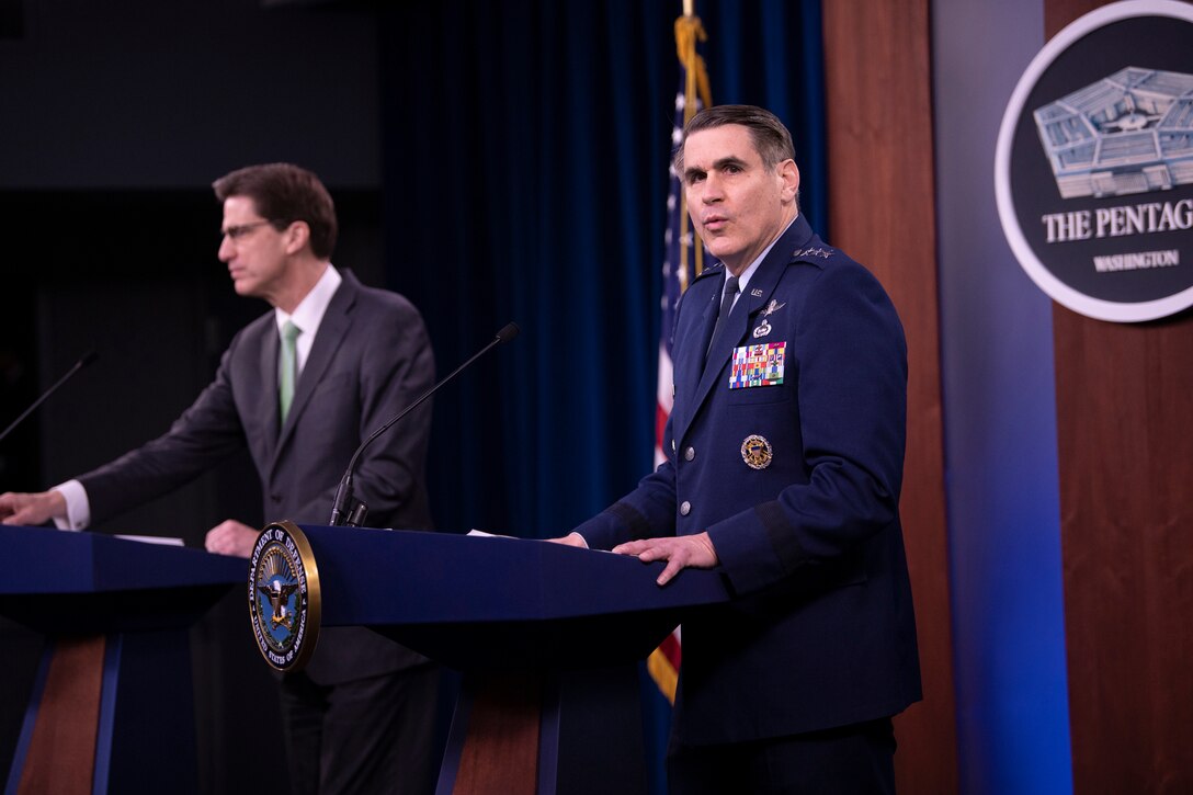 A man in uniform and a man in a business suit stand behind lecterns. Behind them, a sign on the wall says “Pentagon -- Washington.”