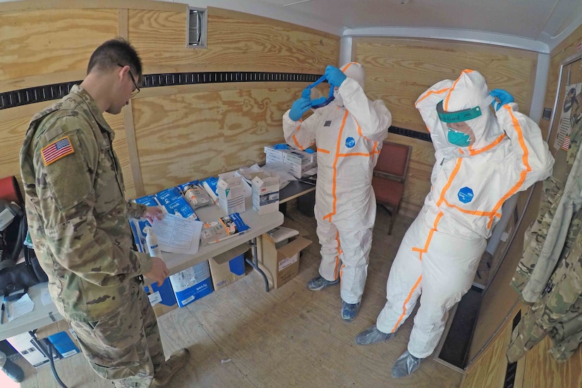 A service member watches two people put on white, protective suits.