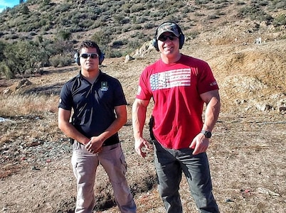 Male in black polo and tan slacks wearing sunglasses and ear protection stands with hands folded in front while standing next to male in red and white tshirt, jeans, ball cap, and hearing protection out in a field.