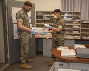Corporal Colten Kokes, S-1 Administration clerk, and Lance Cpl. Zachary Gandiongco, Postal clerk, work together to process necessary mail using mandatory Personal Protective Equipment, such as latex gloves, a protective gaitor for the face or a protective mask, aboard Marine Corps Logistics Base Barstow, Calif., April 8. The new orders for Riverside County, San Bernardino County and MCLB Barstow, require the use of such PPE in public when obtaining necessities, and in circumstances when maintaining the safer six feet away for social distancing.