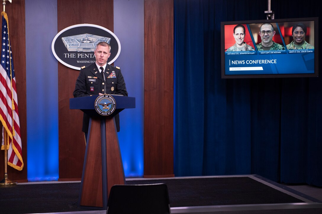 A military officer stands behind a dias with a TV monitor in the background.