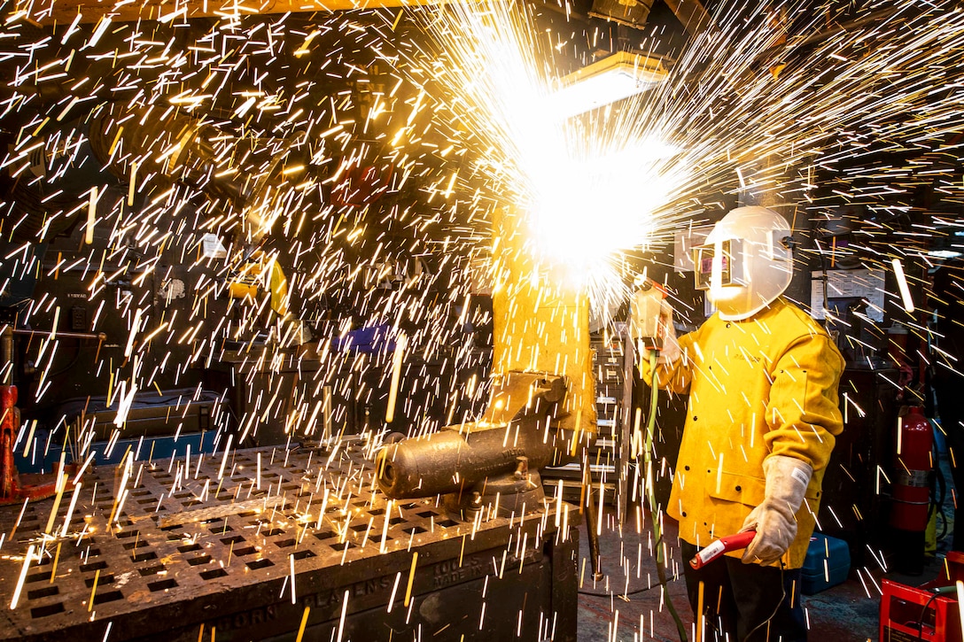 Sparks fly in a room as a sailor in a helmet operates a cutting tool.