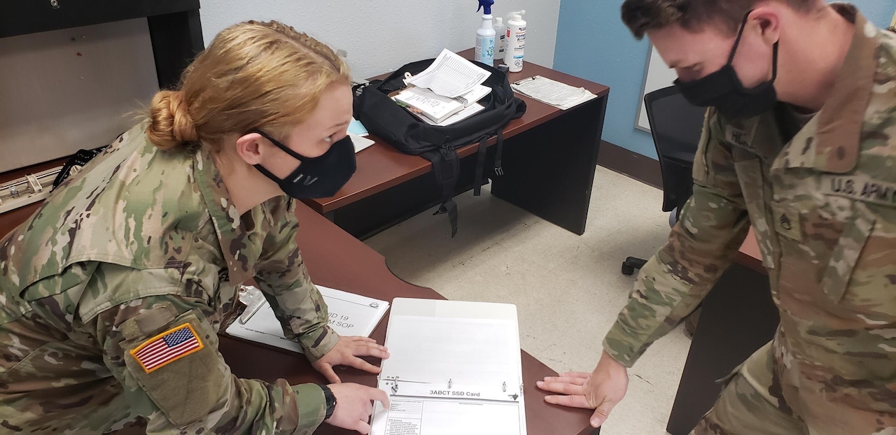 Two soldiers wearing black face masks discuss a report that is in a binder on a desk.