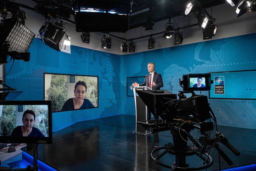 A man stands at lectern and answers questions from a reporter whose face is on television screens to his right and in the foreground.