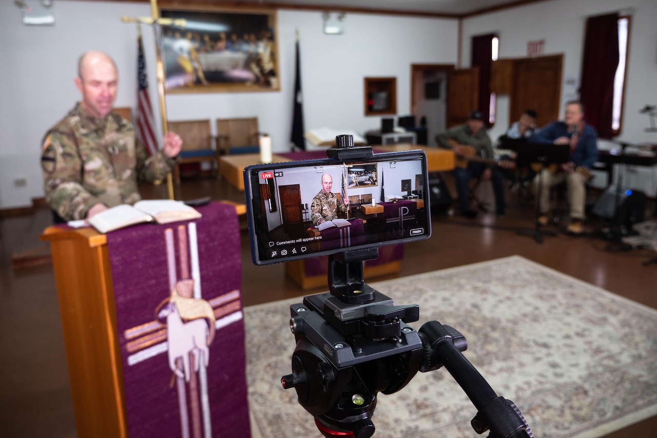 A smartphone in a tripod records a chaplain at a podium as a band sits in the background. A large cross is behind the chaplain.