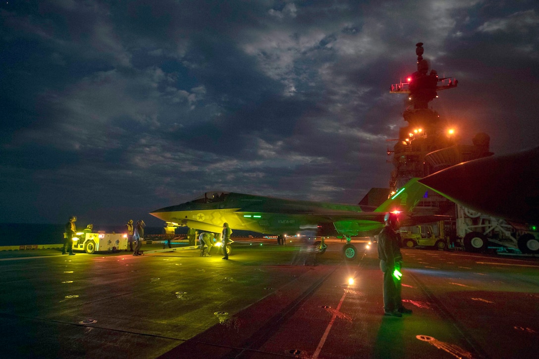 A military aircraft taxis on board a military ship at night.