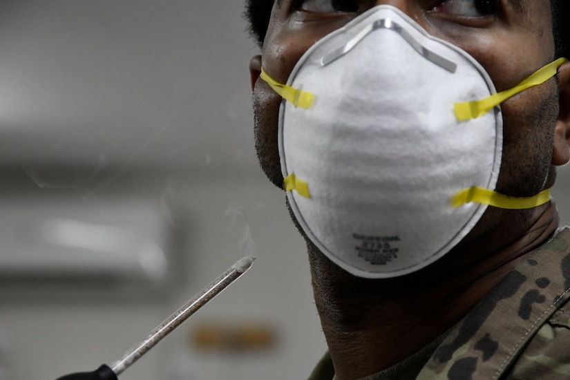 A service member wears a paper mask.  He holds a device up to his face that generates smoke.