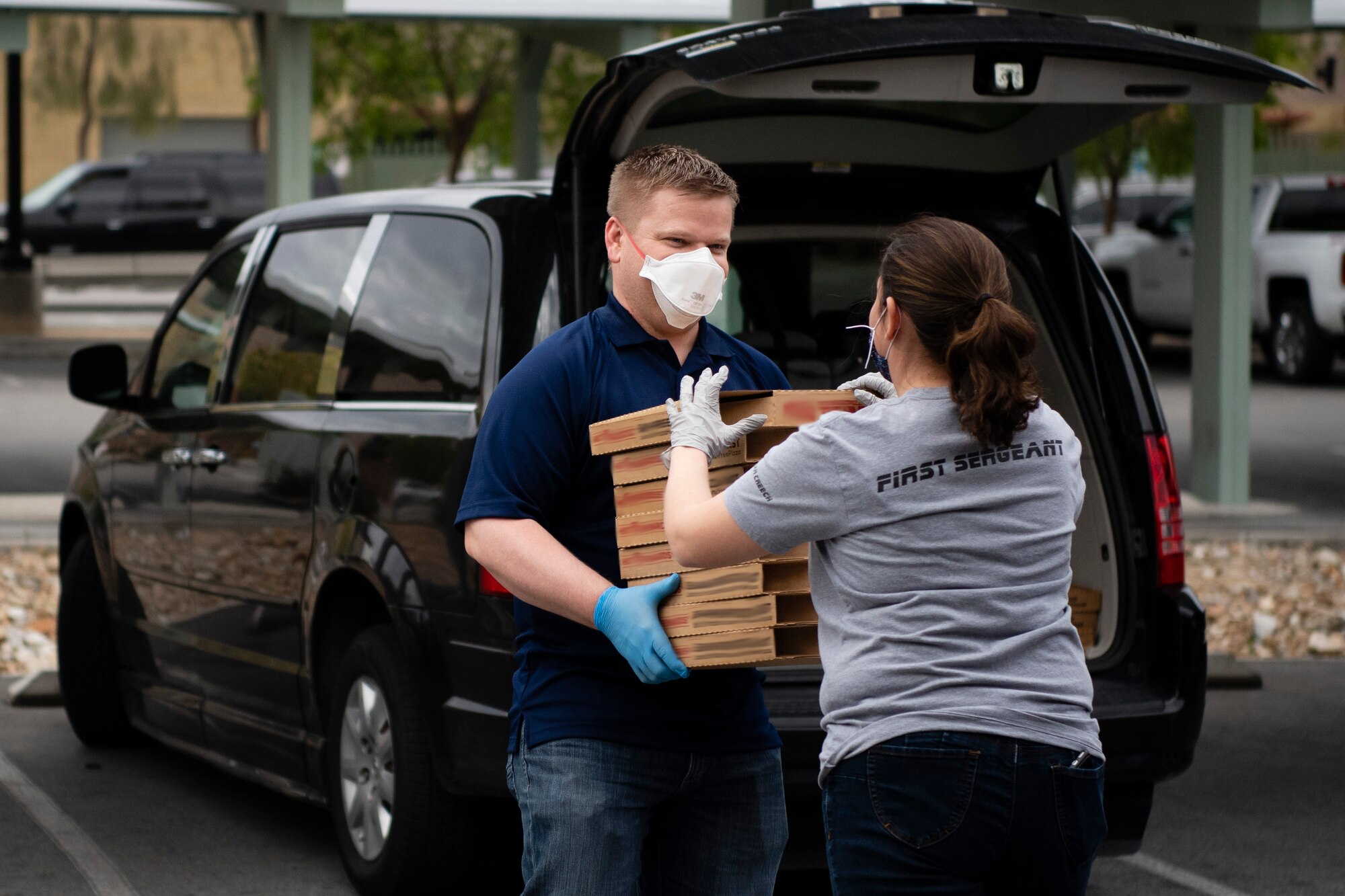 First sergeants, wearing masks and gloves due to the COVID-19 precautions, pass a stack of pizzas to each other in a parking lot.