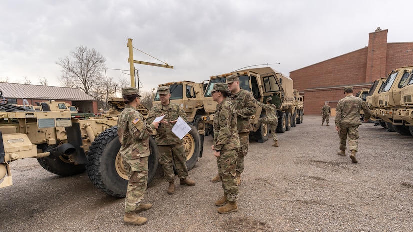 A small group of four troops discuss logistics while other troops board military vehicles in the background.