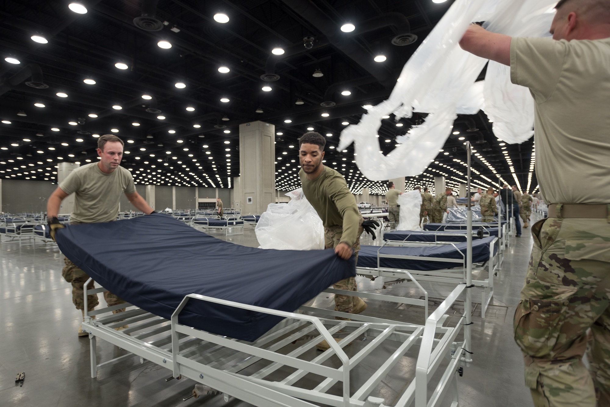 Staff Sgt. Wesley Hack (left) and Senior Airman Trenton Curry (center) of the Kentucky Air National Guard's 123rd Civil Engineer Squadron place a mattress on a hospital bed at the Kentucky Fair and Exposition Center in Louisville, Ky., April 11, 2020. The site, which is expected to be operational April 15, will serve as an Alternate Care Facility for patients suffering from COVID-19 if area hospitals exceed available capacity. The location initially can treat up to 288 patients and is scalable to 2,000 beds. (U.S. Air National Guard photo by Dale Greer)