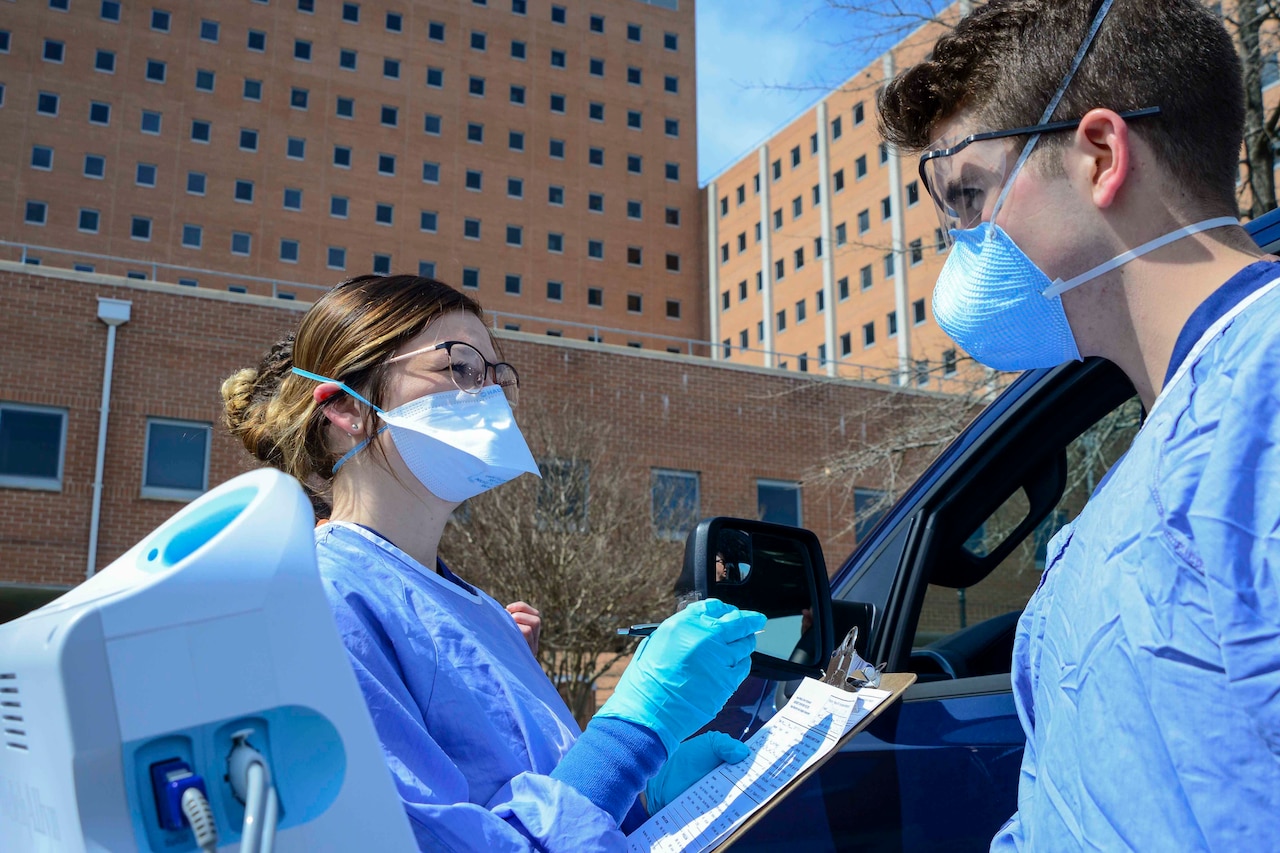 A woman and a man wearing masks and other protective gear talk as the woman writes on a clipboard.