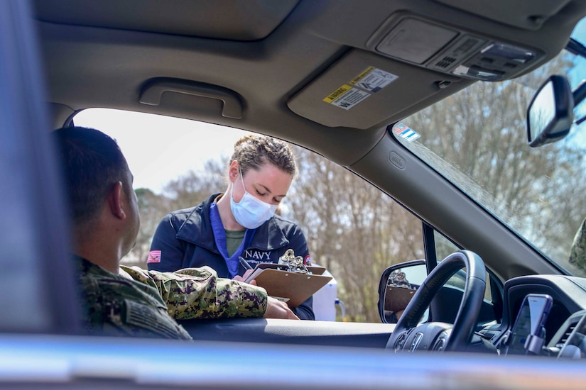 A woman wearing a protective mask and writing on a clipboard checks in a driver at drive-thru screening checkpoint.