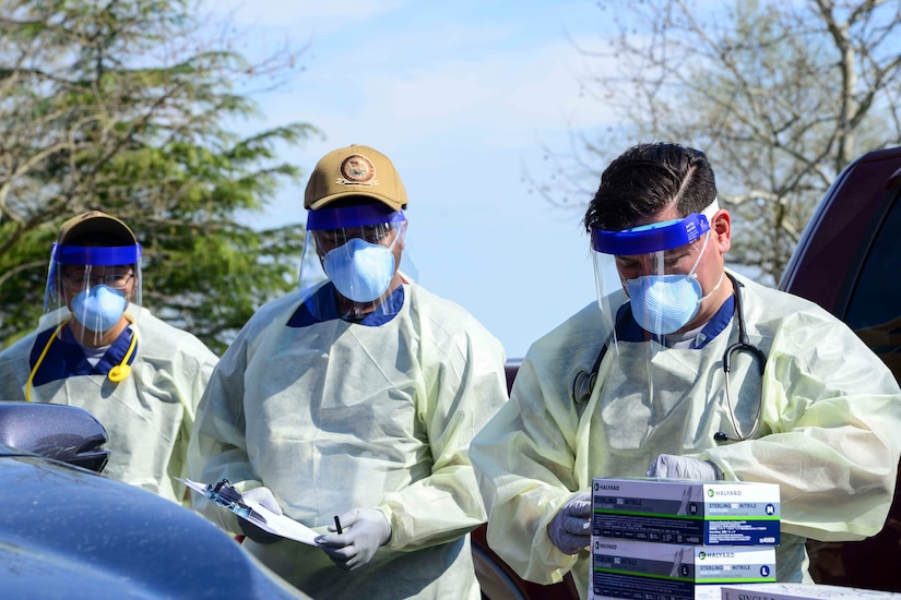 Three men wearing masks, face shields and other protective equipment screen a patient at a drive-thru screening checkpoint.