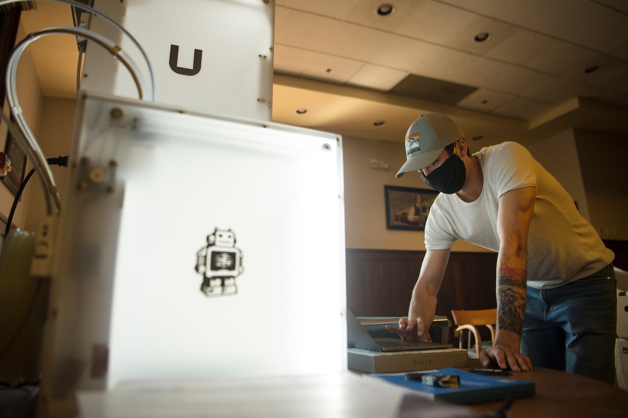 An airman wearing a black mask and baseball cap works on a 3D printer.
