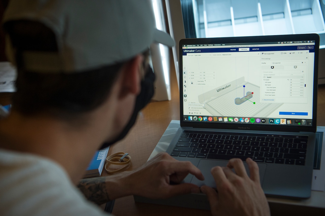 View over the shoulder of an airman as he looks at a laptop monitor while operating a 3D printer.