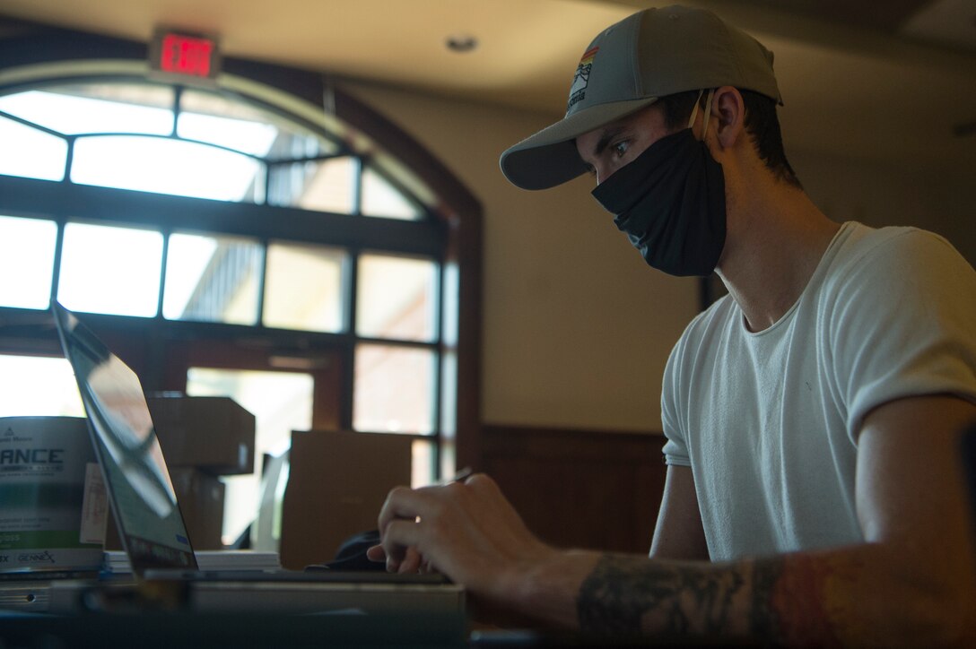 An airman wearing a black face mask works on a laptop computer.