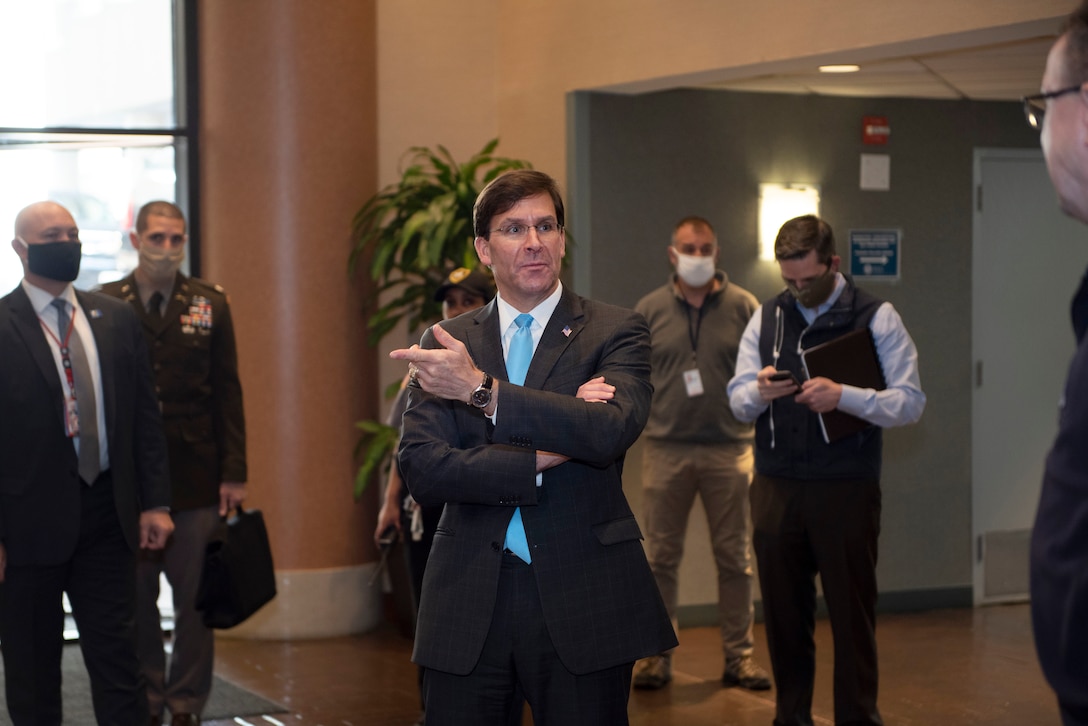 Defense Secretary Dr. Mark T. Esper stands in a lobby-type area with several people wearing masks.