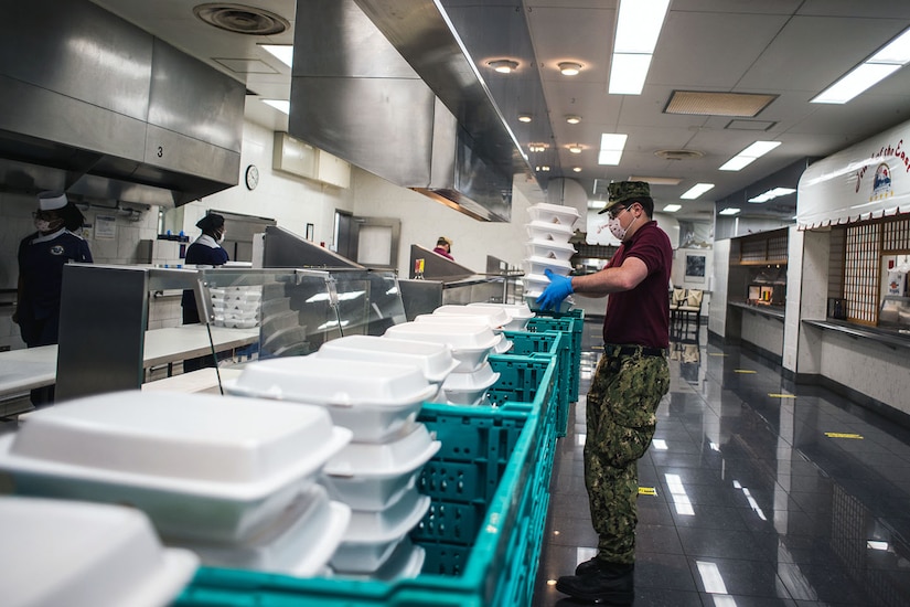 A sailor holds a stack of food containers while standing by crates filled with other containers in a cafeteria kitchen.