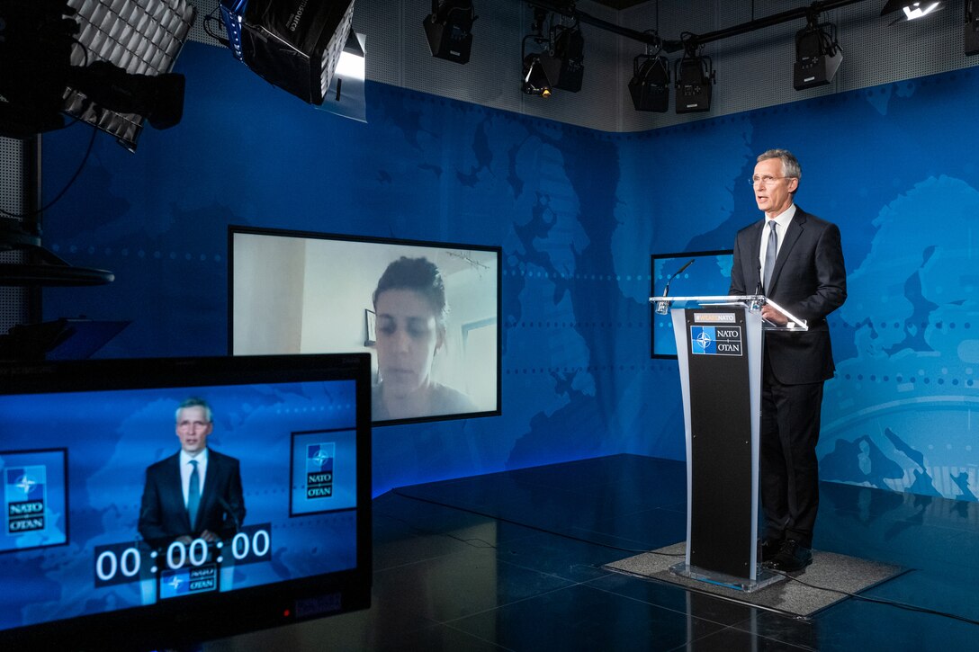 A man stands behind a lectern at a virtual meeting while a remotely located participant shows on a nearby screen.