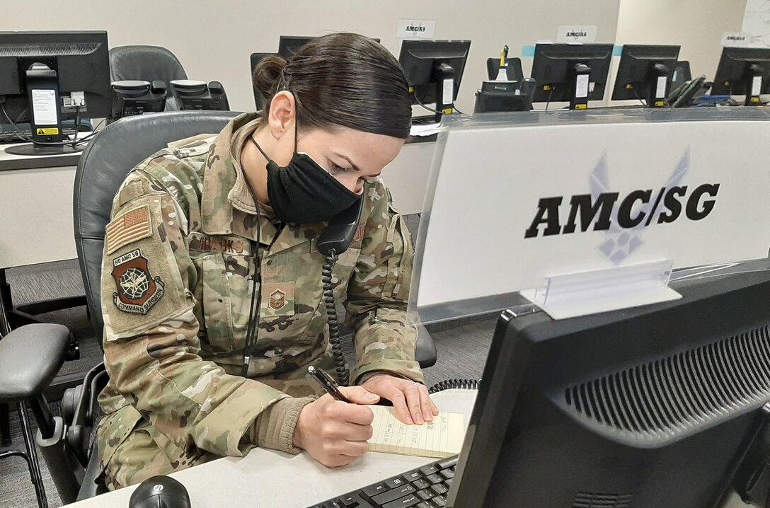 An airman wearing a black face mask and working at a computer station marked “AMC/SG” writes in a notepad.