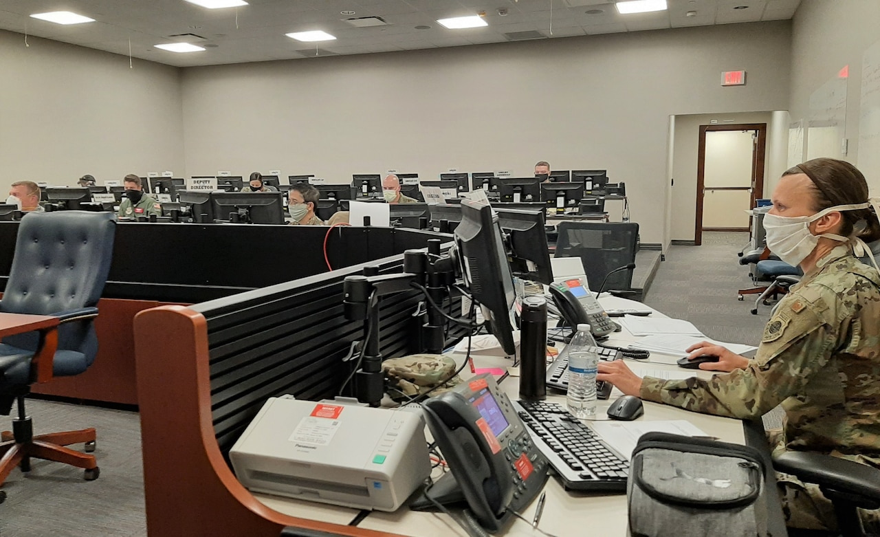 An Air Force officer wearing a white face mask works at a computer station in a large room with others working at their computer stations in the background.
