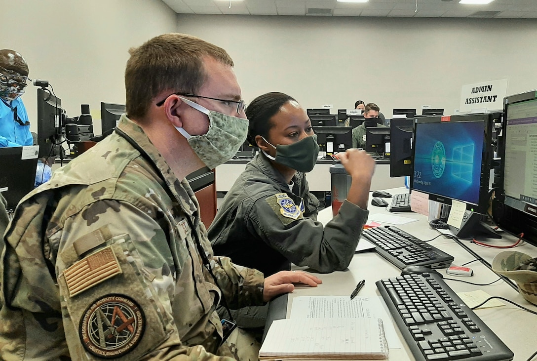 Two airmen wearing face masks talk while working at a computer station.