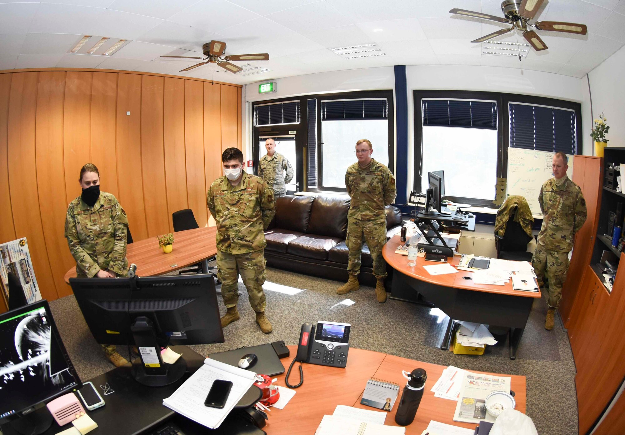 Five Airmen stand in an office, looking at a computer monitor during a teleconference call