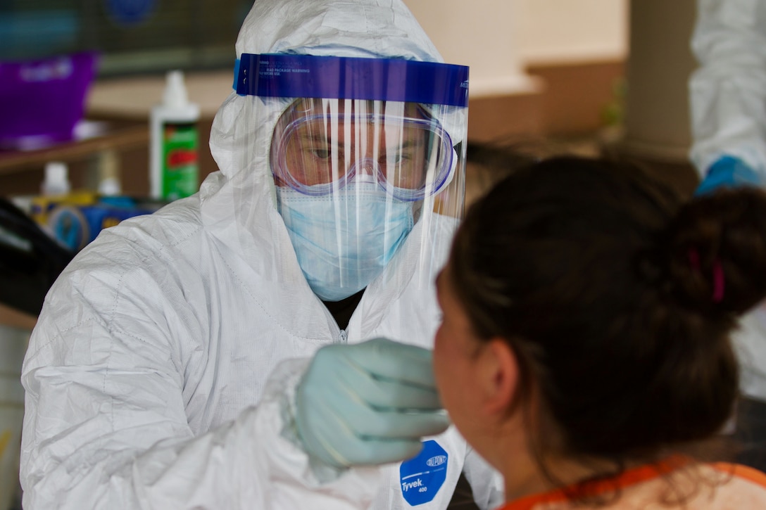 A soldier wearing full protective gear reaches toward a woman’s face to obtain a sample for COVID-19 testing.