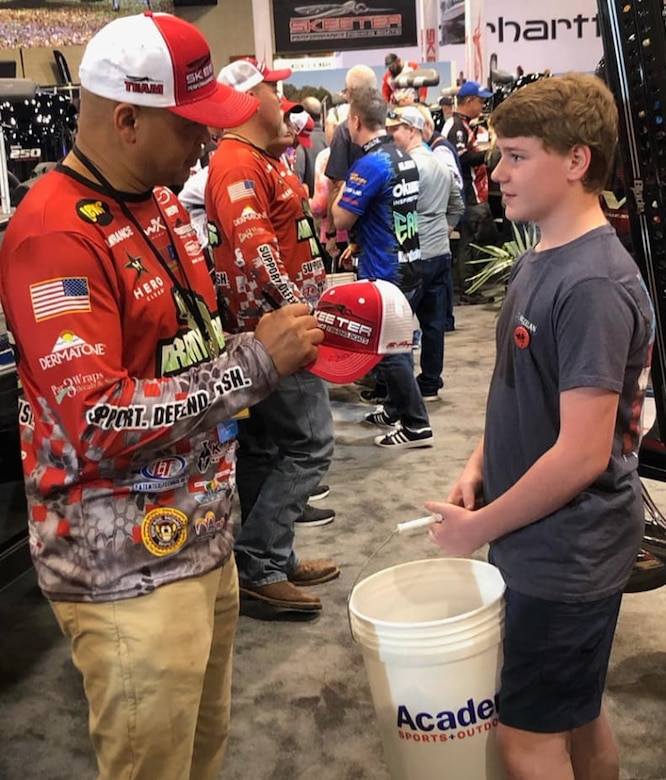 white mail in red multi colored printed shirt with red and white hat holds marker and hat while with young boy stands holding a white bucket