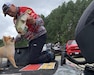 Man in bright red printed shirt and black pants and black and white cap holds a fish over a small Pomeranian dog.