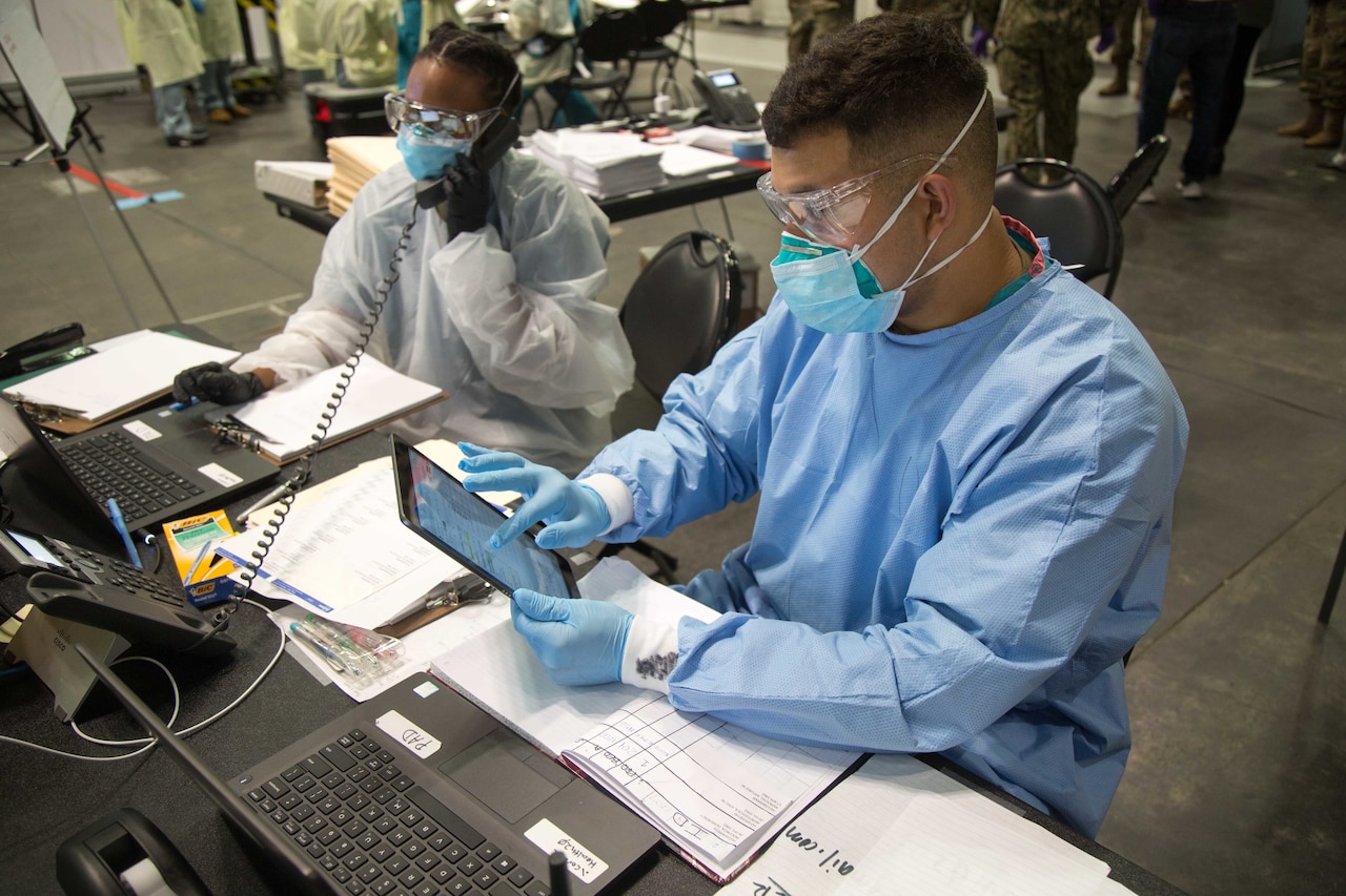 One soldier speaks on a telephone and another uses a hand-held tablet. Both are wearing face masks and other protective gear and are seated at laptop computers.