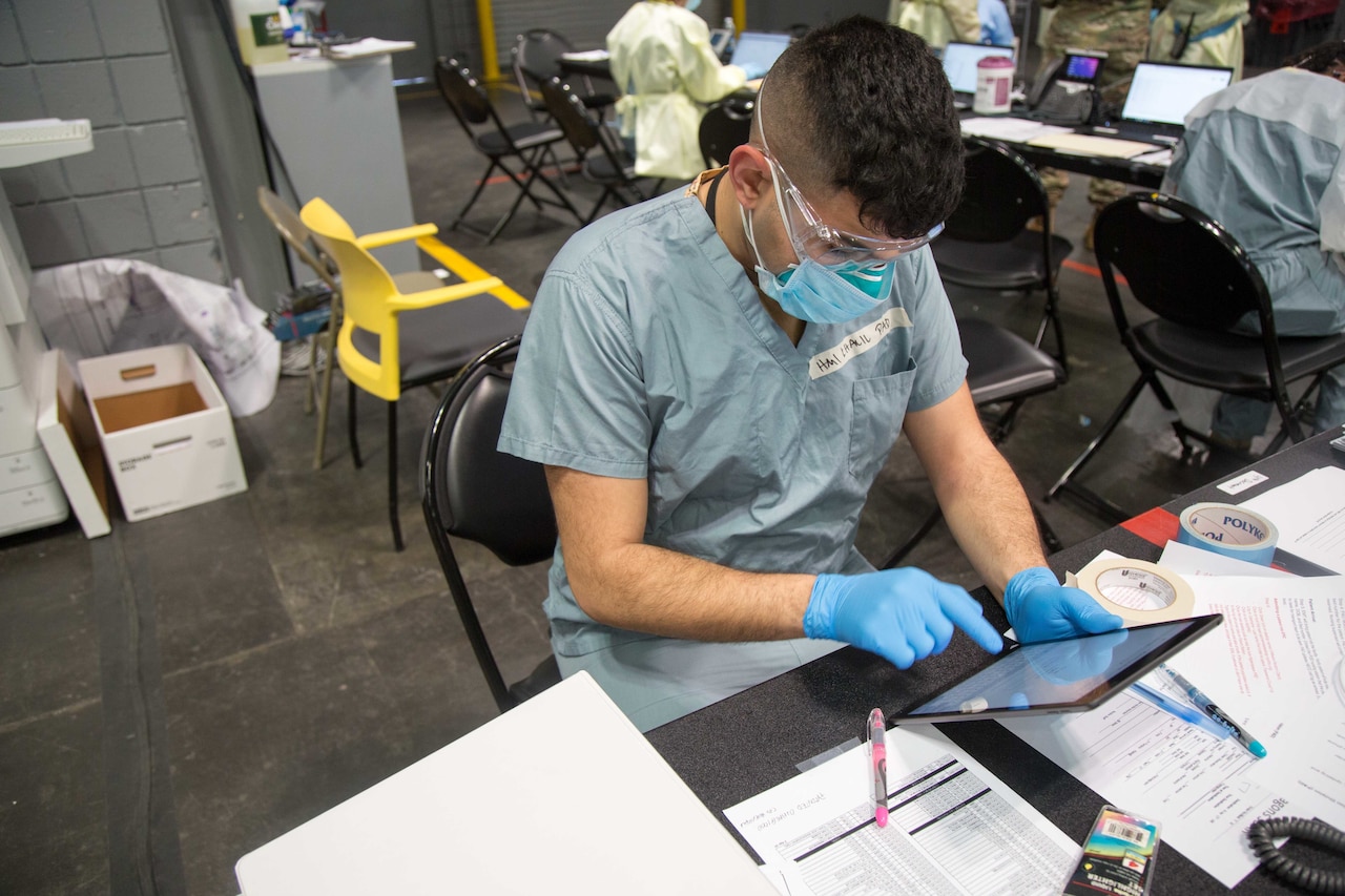 A sailor wearing a mask and other protective gear works with a hand-held tablet while seated at a table.