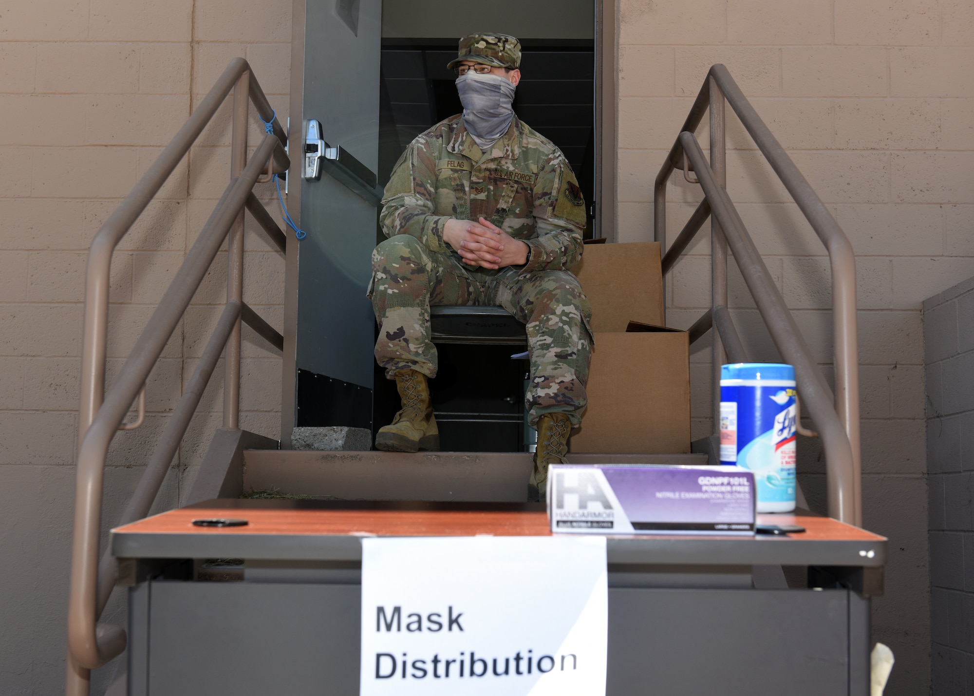 A photo of a U.S. Airman sitting on doorsteps waiting to distribute cloth masks