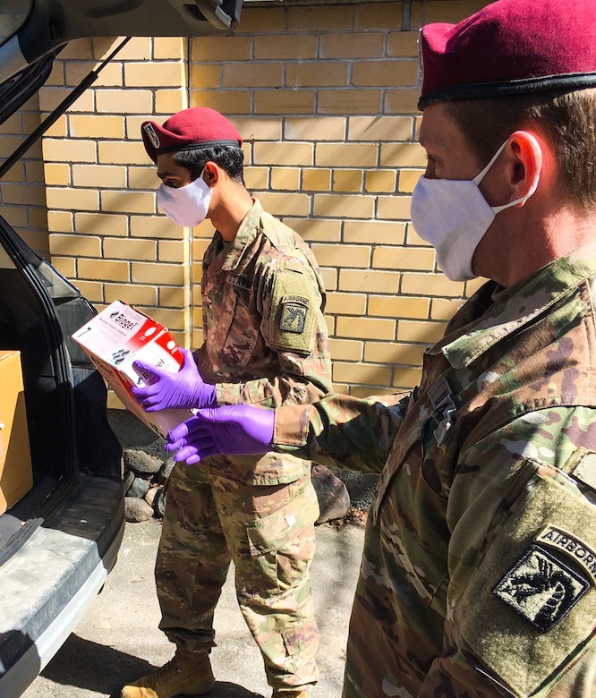 Two uniformed service members wearing face masks and gloves unload medical gear from the back of a vehicle.