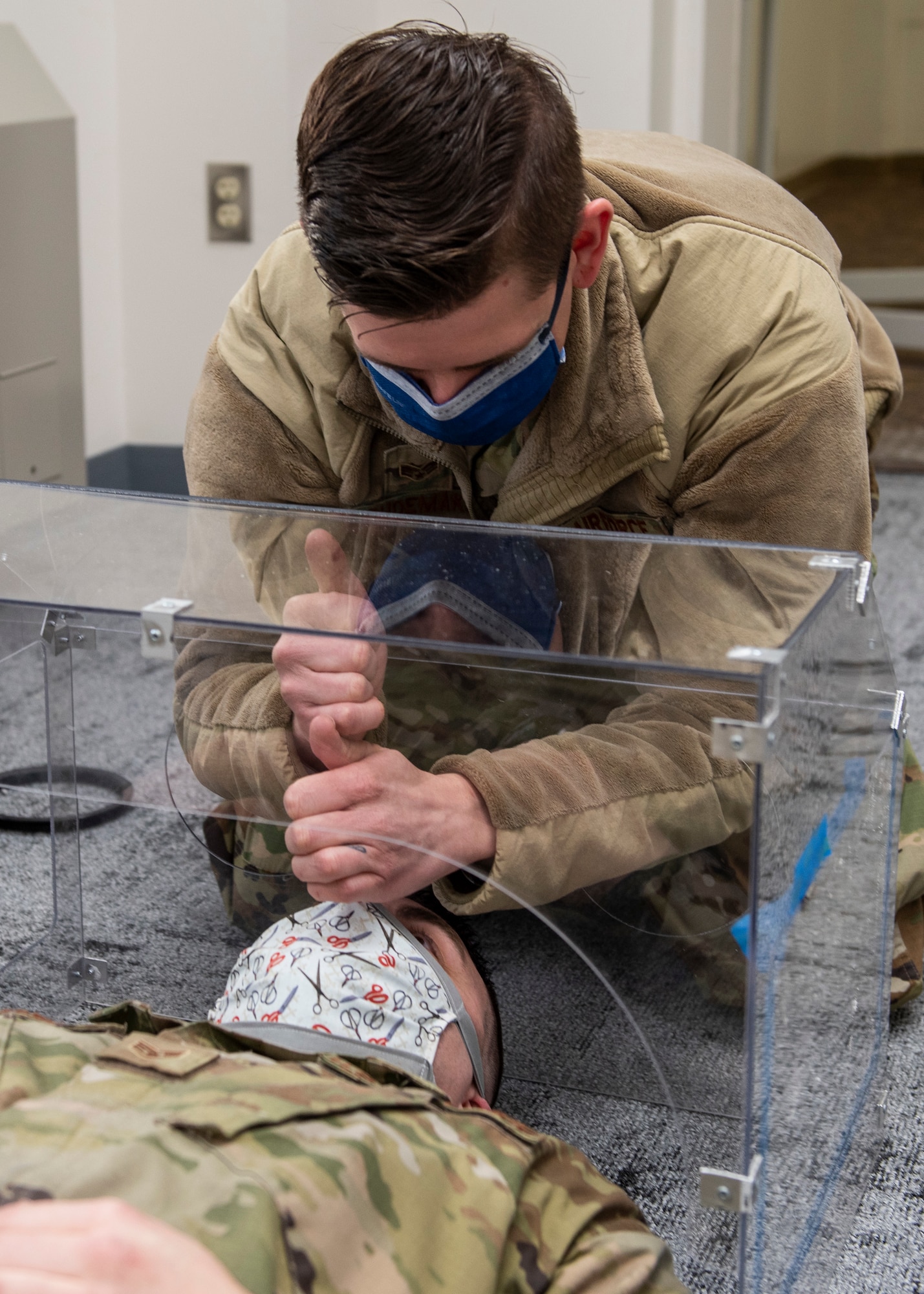 U.S. Air Force Senior Airman Michael Shoemaker, 673d Medical Support Squadron biomedical equipment technician, demonstrates how intubation might look on U.S. Air Force Staff Sgt. Andrew Taylor, 673d Medical Support Squadron medical logistics noncommissioned officer in charge of acquisitions, under an innovated, plastic polycarbonate enclosure at Joint Base Elmendorf-Richardson, Alaska, April 7, 2020. Shoemaker and Taylor designed and built the reusable device to be a protective barrier between medical providers and a patient to prevent possible exposure to COVID-19 and other airborne diseases. The enclosure can be placed over a patient’s head and upper torso prior to intubation or similar treatments, with access to the patient via two holes at the head of the enclosure for a physician's hands and arms, and two side doors for additional access.