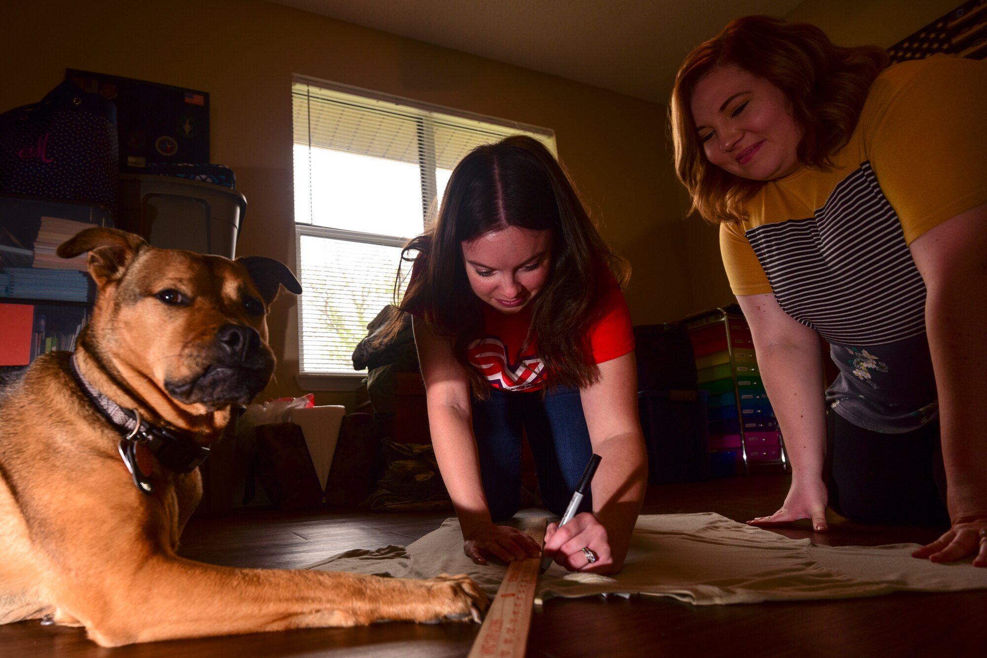 Accompanied by canine companion, Sadie, Mallory Albers, Science Technology Engineering Mathematics Magnet school special education teacher at Laughlin Air Force Base, Texas, and Chelsea Truster, St. James Episcopal Pre-K teacher and Laughlin spouse, measure out patterns for face masks to help protect from COVID-19 on a uniform on base, April 10, 2020. Learning through trial and error, they work on perfecting their process for making face masks to match Airmen’s uniforms. (U.S. Air Force Photo by Senior Airman Anne McCready)