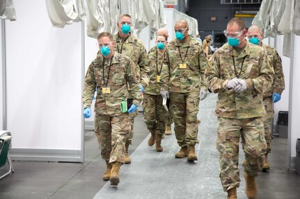 Six soldiers wearing blue face masks walk through the patient care area of an alternate care facility set up as part of the COVID-19 pandemic response.