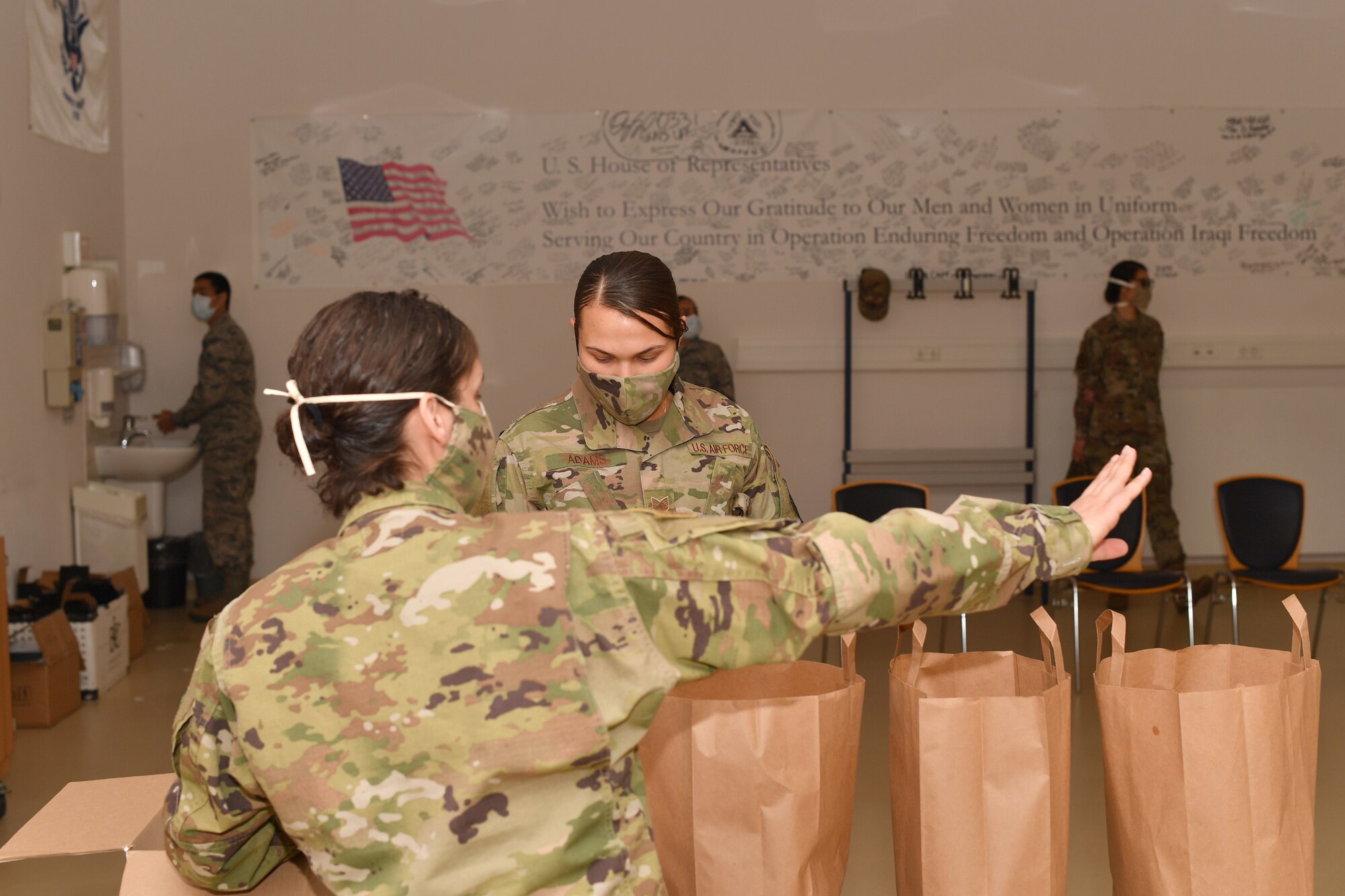 An Airman directs another Airman down a line of to-go meals, while others stand in line and wash their hands.