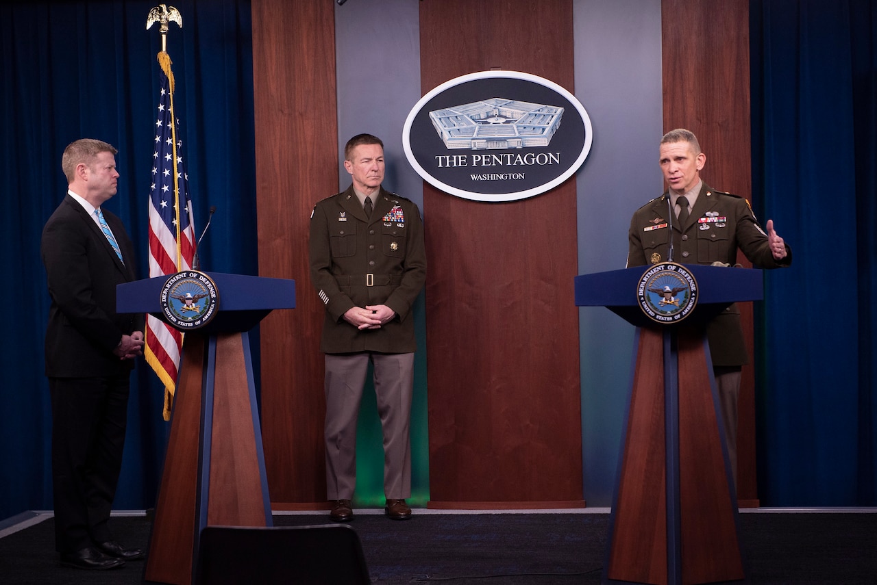 A senior enlisted soldier speaks at a lectern as an officer in uniform and a man wearing a suit stand on the same stage.