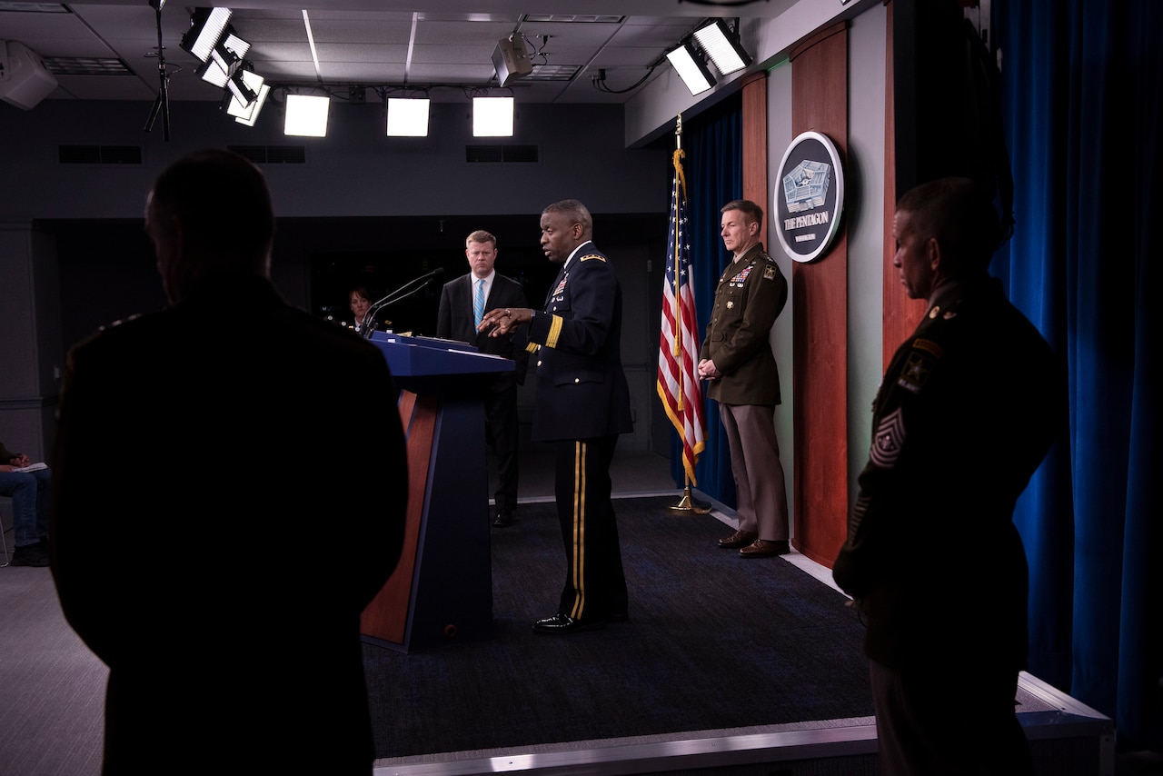Side view of an Army officer standing at a lectern with other people in the foreground and background.