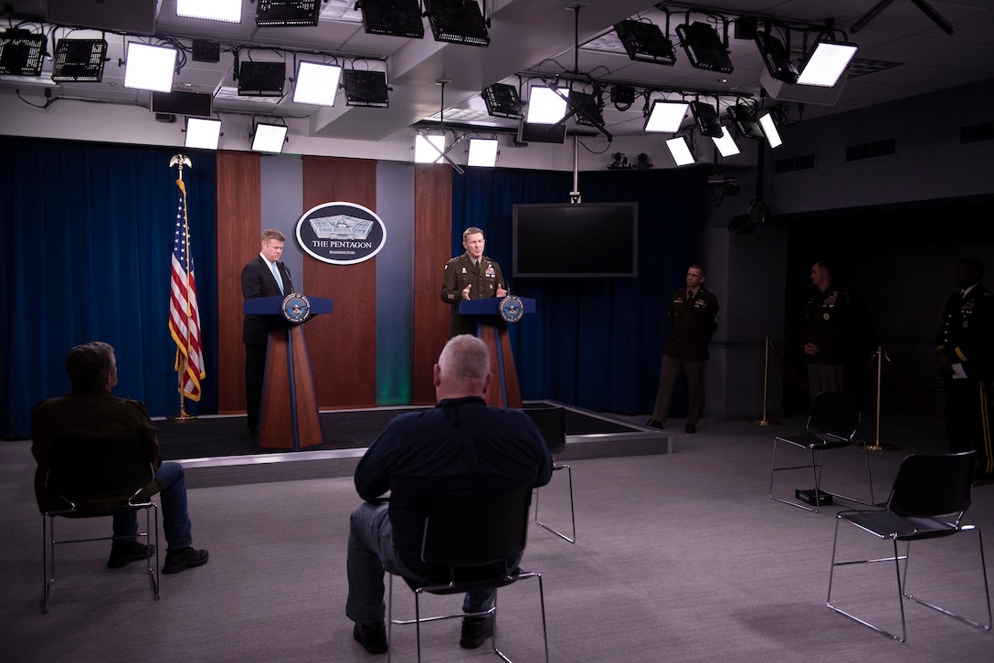 A man wearing a suit and an Army officer in uniform stand at separate lecterns as they brief reporters who are seated in observance of social distancing guidelines.
